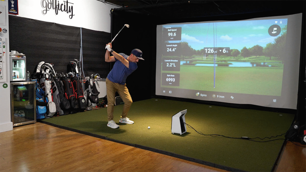 Golfer mid-swing using a Garmin Approach R50 radar launch monitor with ball flight data displayed on simulator screen